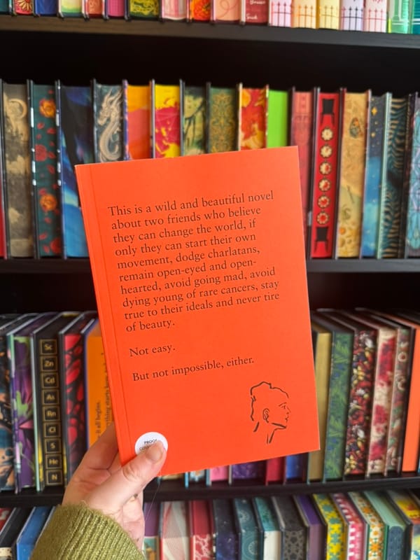 A hand holds the bright orange proof copy of a book in front of a bookshelf filled with colourful special edition hardbacks. 