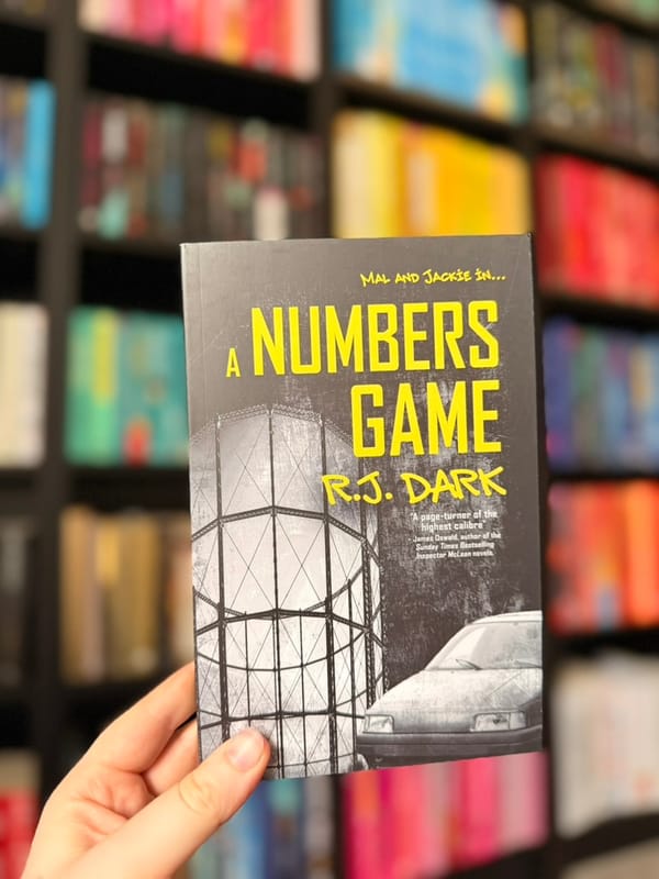 A hand holds a copy of A Numbers Game by R.J. Dark in front of softly blurred rainbow bookshelves. 