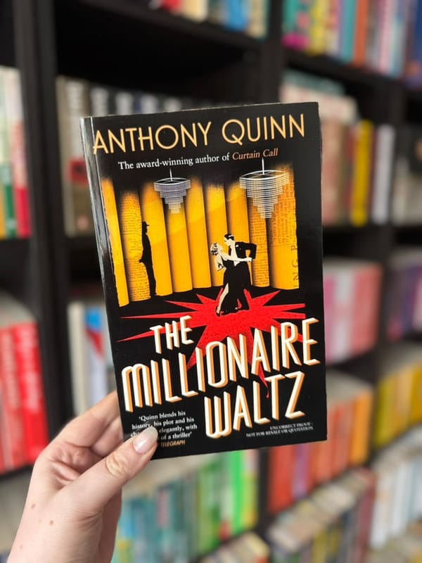 A hand holds "The Millionaire Waltz" book by Anthony Quinn, featuring a couple dancing on stage. Bookshelf background, colorful spines.