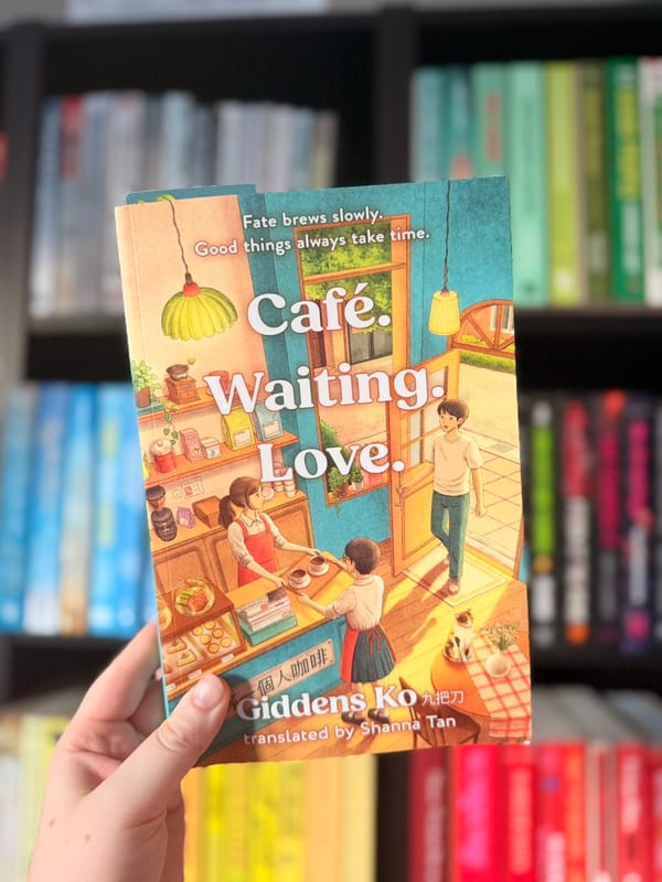 A hand holds a copy of Café. Waiting. Love. by Giddens Ko, translated by Shanna Tan, in front of a softly blurred, colour-coordinated bookshelf.