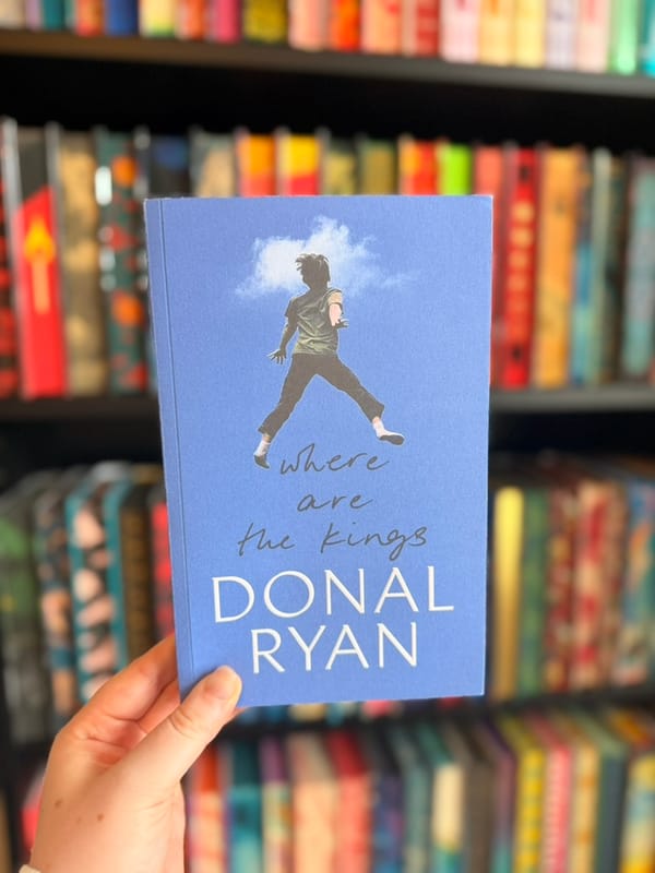 A hand holds a copy of Where Are the Kings by Donal Ryan in front of a softly blurred bookshelf filled with colourful books. 
