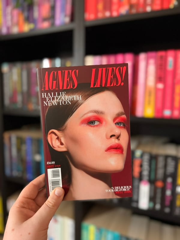 A hand holds a copy of Agnes, Lives! by Hallie Elizabeth Newton in front of softly blurred rainbow bookshelves.