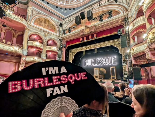 Interior of the Alhambra Theatre with the Burlesque stage in the background and a black fan in the foreground reading “I’m a Burlesque Fan"