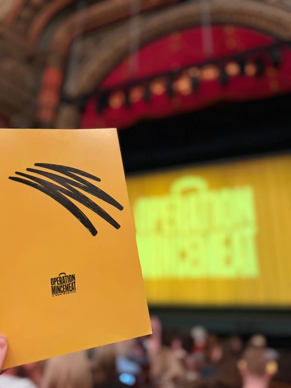 yellow Operation Mincemeat programme held up inside the theatre, with the brightly lit stage and audience blurred in the background.